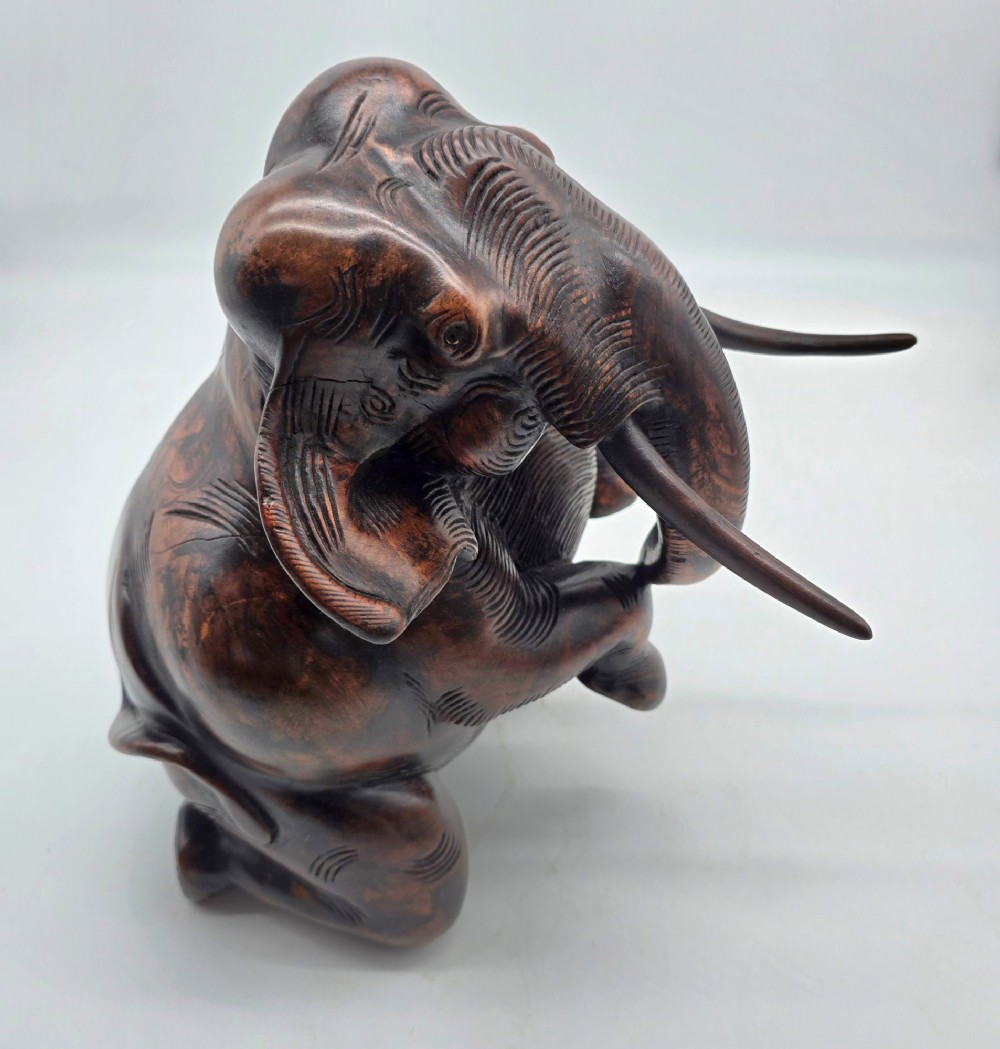 A dark carved hardwood sculpture of an elephant seated on its hind legs with its trunk raised and long tusks exposed. View X