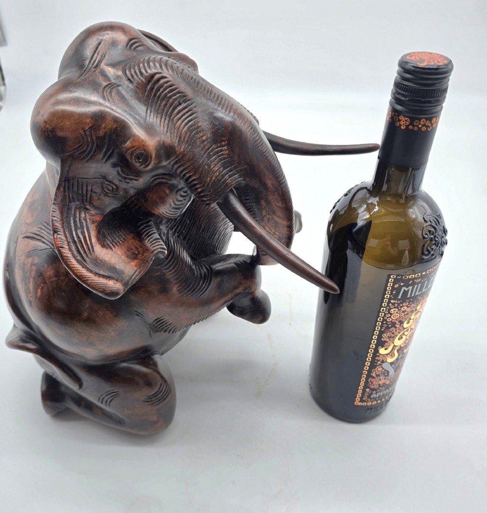 A dark carved hardwood sculpture of an elephant seated on its hind legs with its trunk raised and long tusks exposed. View XI