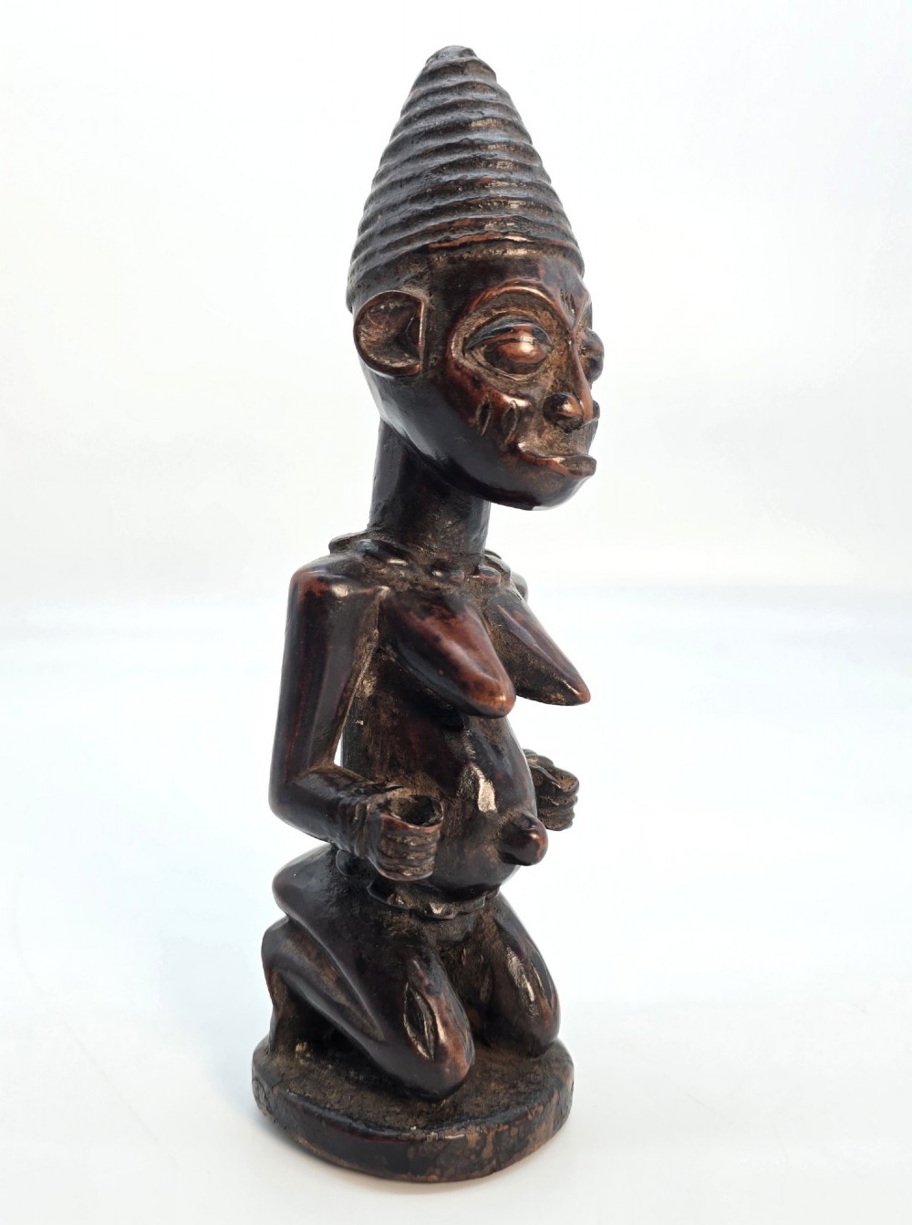 A dark brown hand-carved wooden figure depicting a kneeling female with a tall ridged headpiece and carved facial markings.