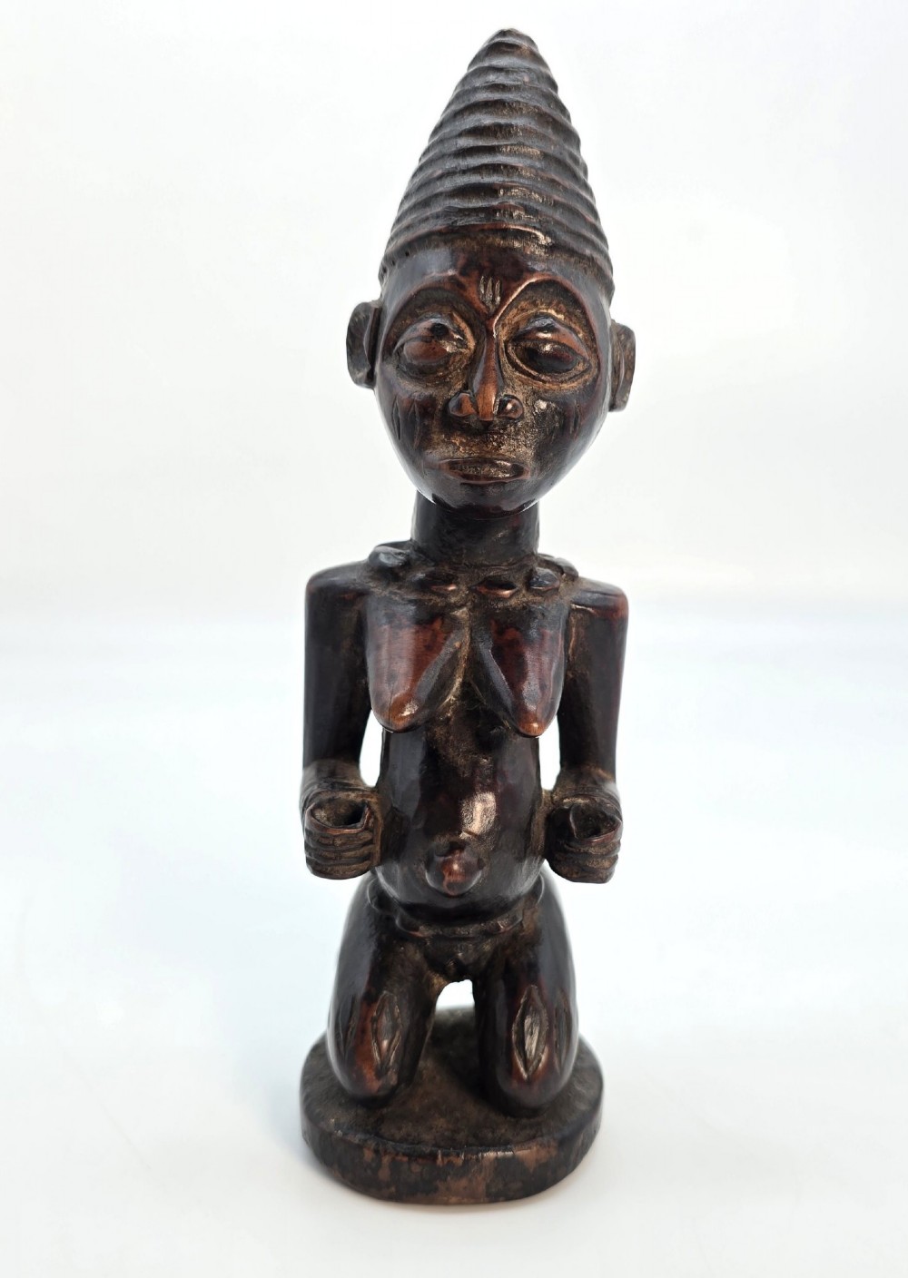 A dark brown hand-carved wooden figure depicting a kneeling female with a tall ridged headpiece and carved facial markings. View II