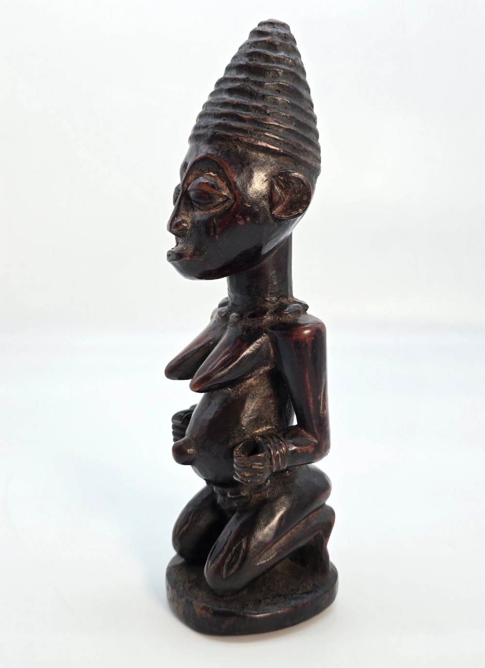 A dark brown hand-carved wooden figure depicting a kneeling female with a tall ridged headpiece and carved facial markings. View III