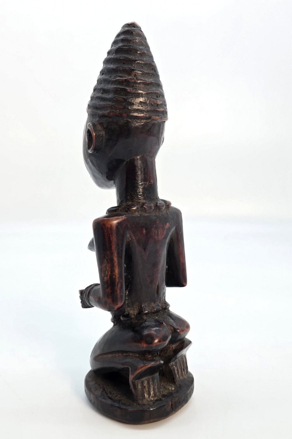 A dark brown hand-carved wooden figure depicting a kneeling female with a tall ridged headpiece and carved facial markings. View IV