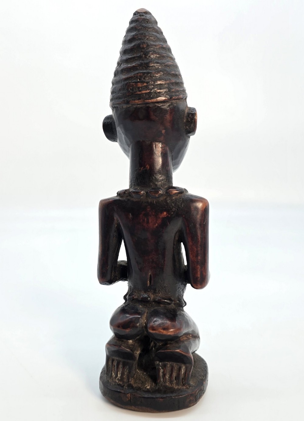 A dark brown hand-carved wooden figure depicting a kneeling female with a tall ridged headpiece and carved facial markings. View V