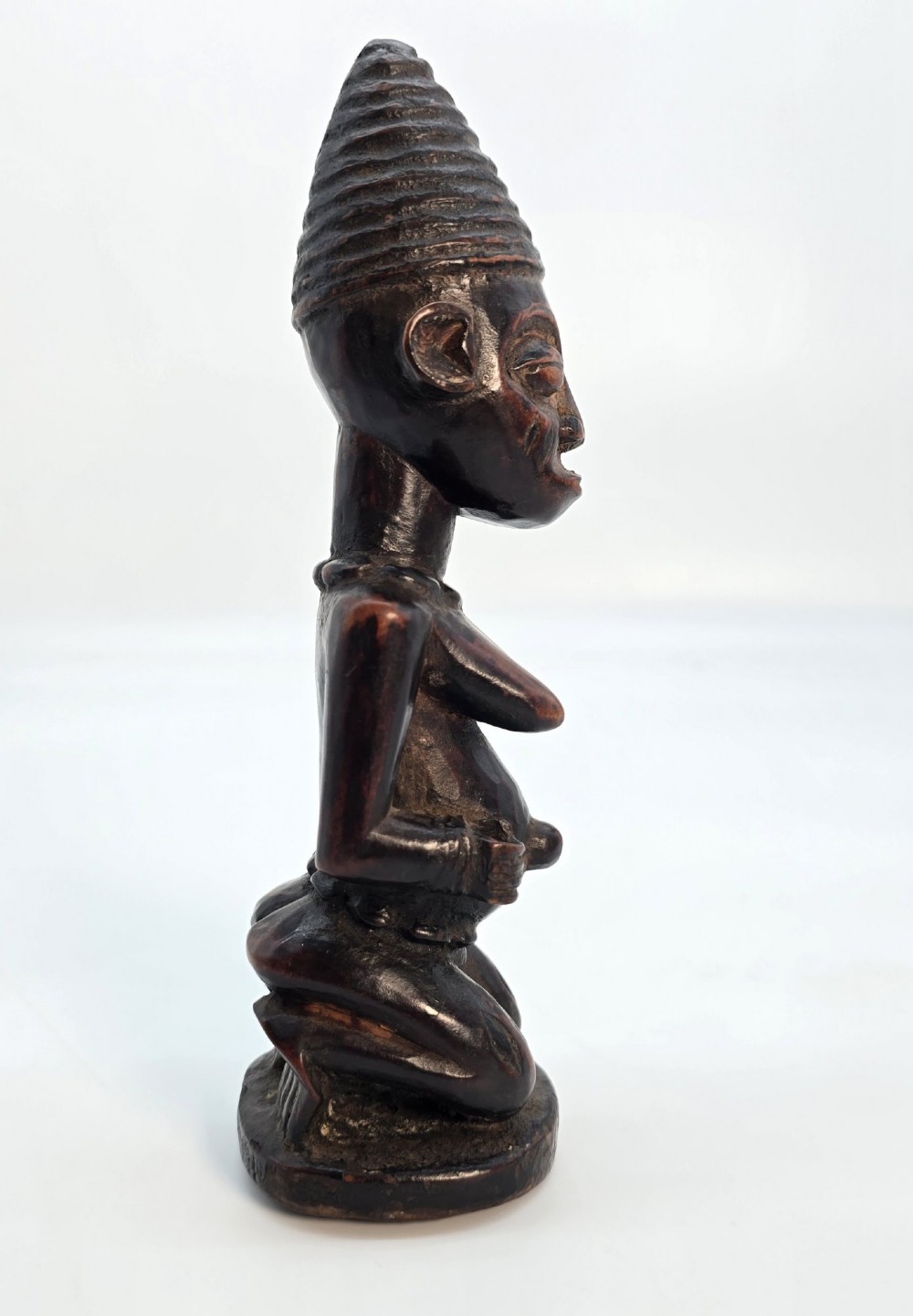 A dark brown hand-carved wooden figure depicting a kneeling female with a tall ridged headpiece and carved facial markings. View VI
