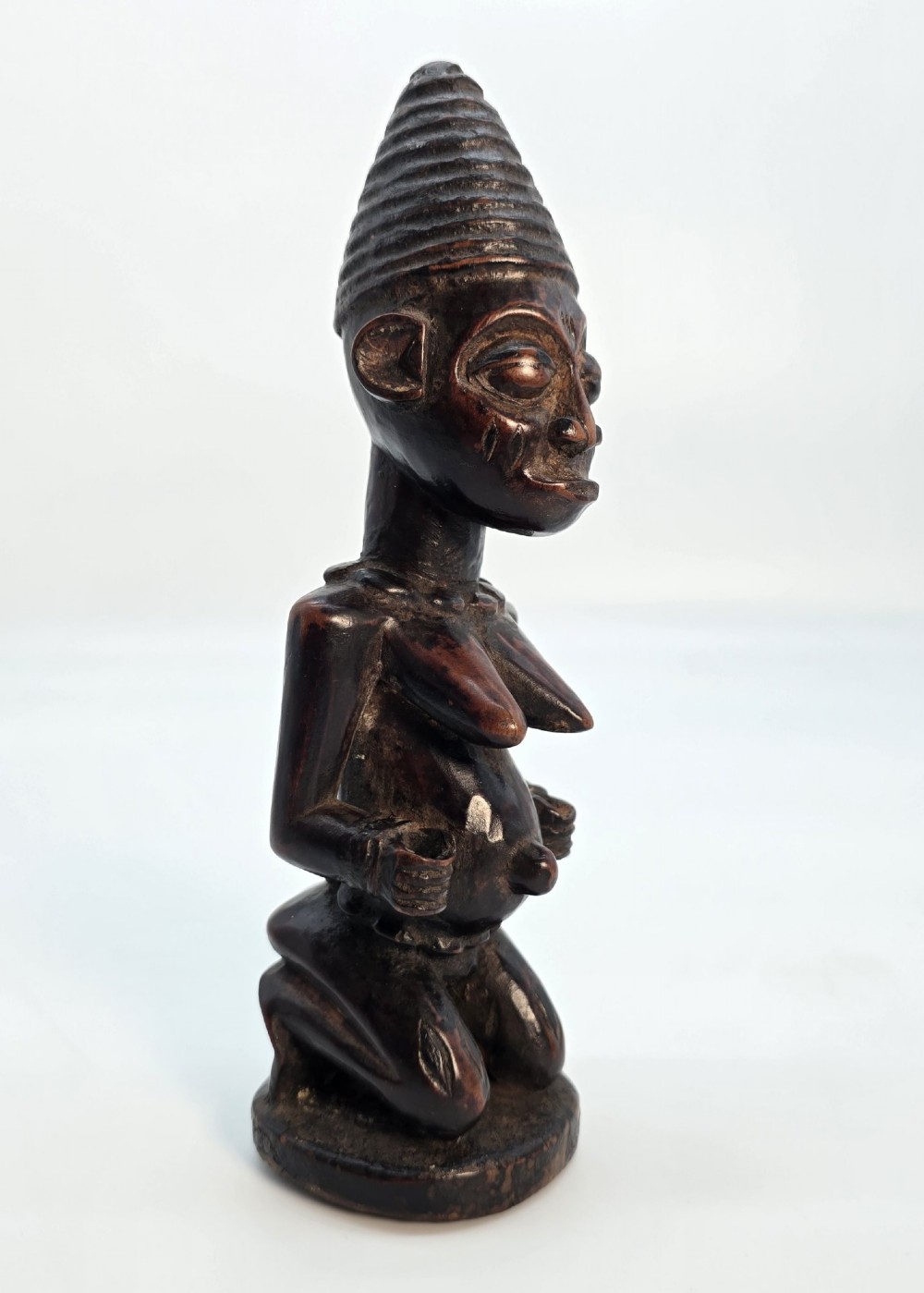 A dark brown hand-carved wooden figure depicting a kneeling female with a tall ridged headpiece and carved facial markings. View VII