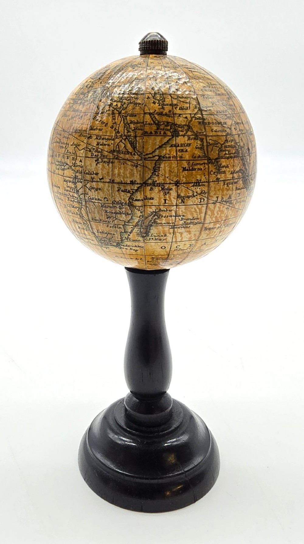 A miniature terrestrial globe with printed sepia paper gores resting on a turned black ebonised wooden pedestal stand with a metal top finial. View II