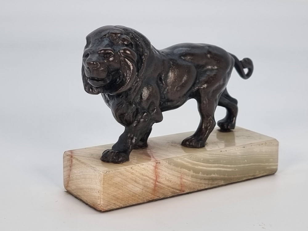 A dark patinated bronze sculpture of a prowling lion mounted on a rectangular cream veined onyx base. View II