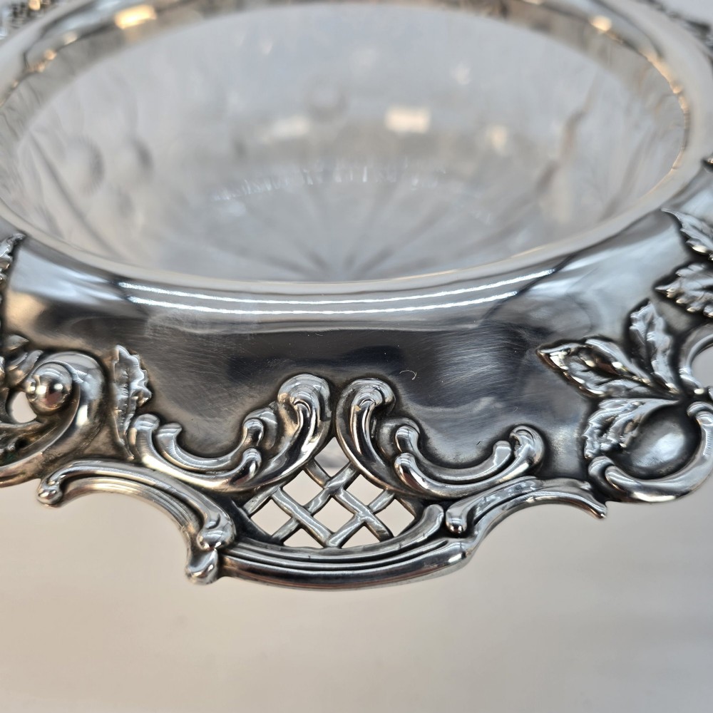 A clear crystal bowl featuring an intricately chased sterling silver rim decorated with floral motifs, scrolls, and lattice patterns. View X