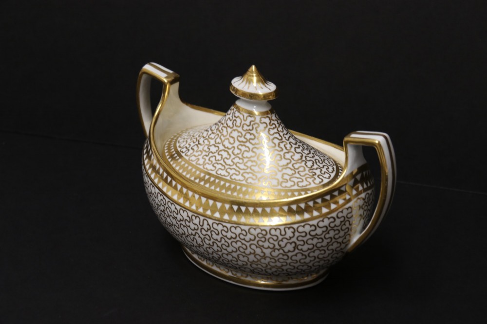 An antique white porcelain covered sucrier decorated with intricate gold scroll patterning, geometric trim, twin loop handles, and a pointed finial lid. View II