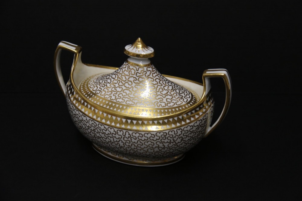 An antique white porcelain covered sucrier decorated with intricate gold scroll patterning, geometric trim, twin loop handles, and a pointed finial lid. View III