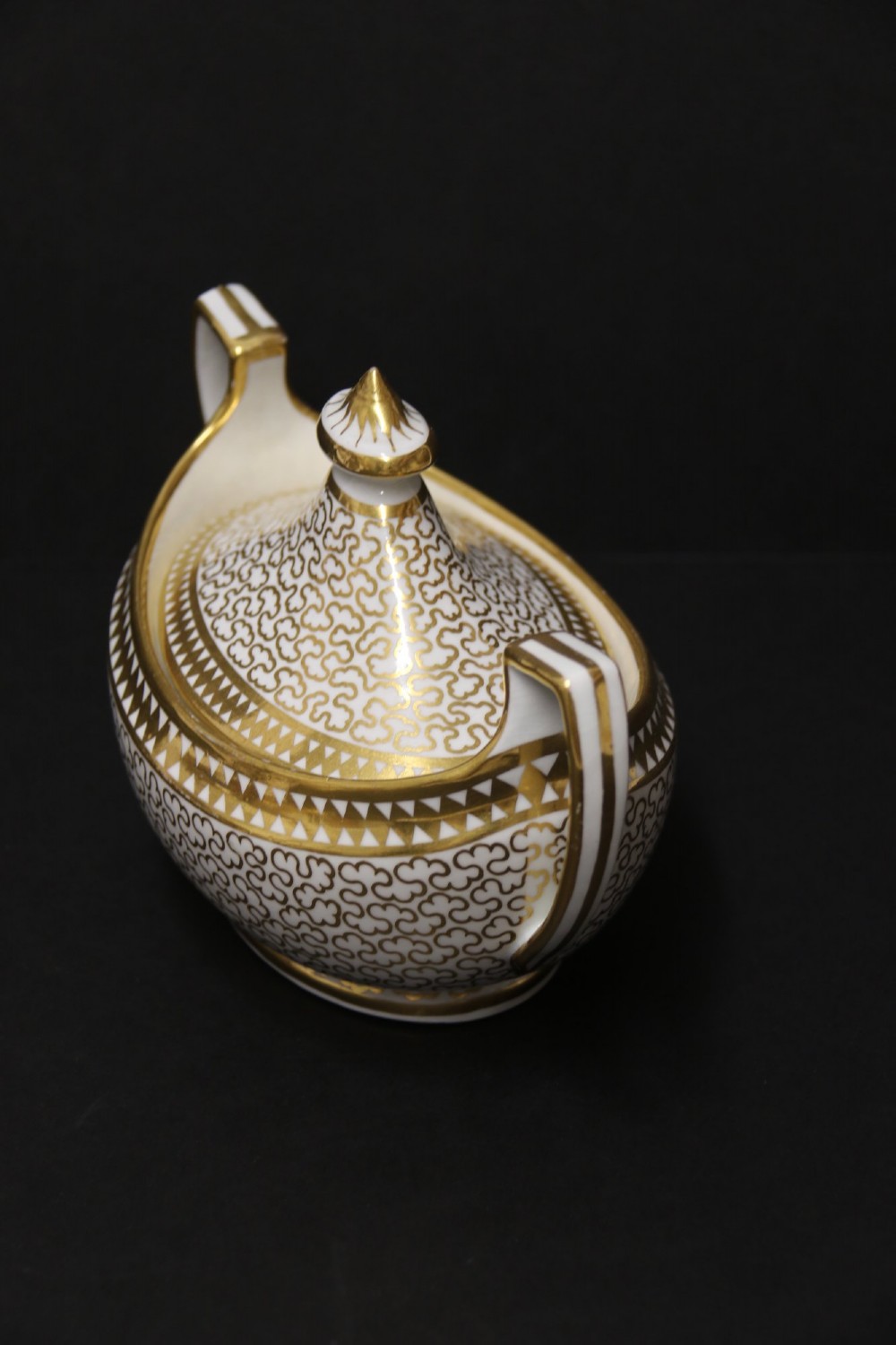 An antique white porcelain covered sucrier decorated with intricate gold scroll patterning, geometric trim, twin loop handles, and a pointed finial lid. View IV