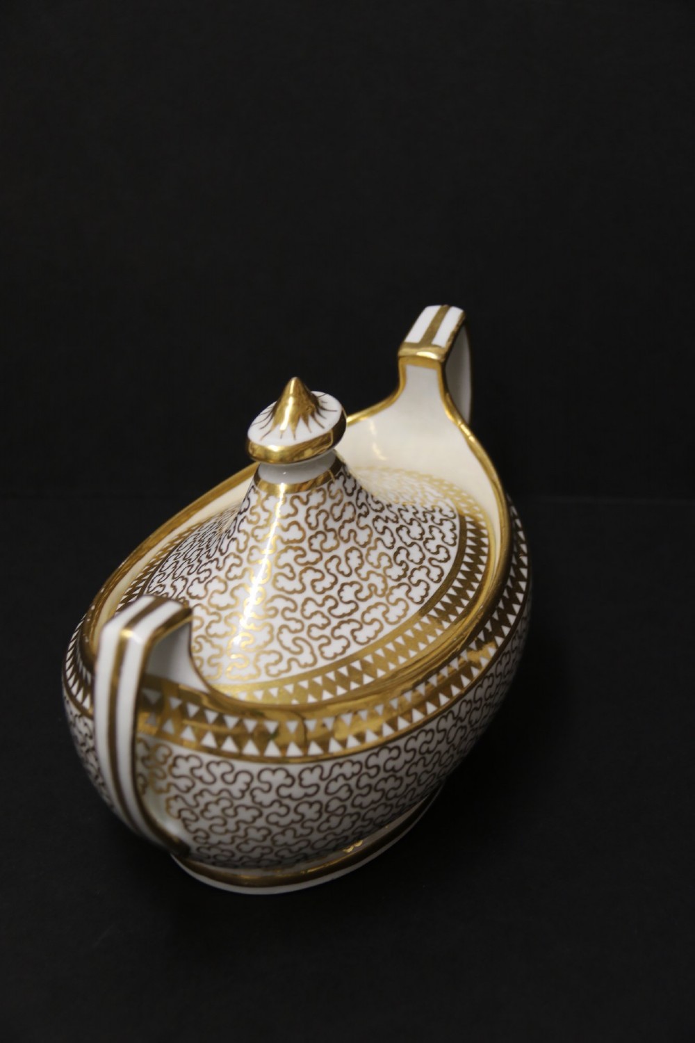 An antique white porcelain covered sucrier decorated with intricate gold scroll patterning, geometric trim, twin loop handles, and a pointed finial lid. View V