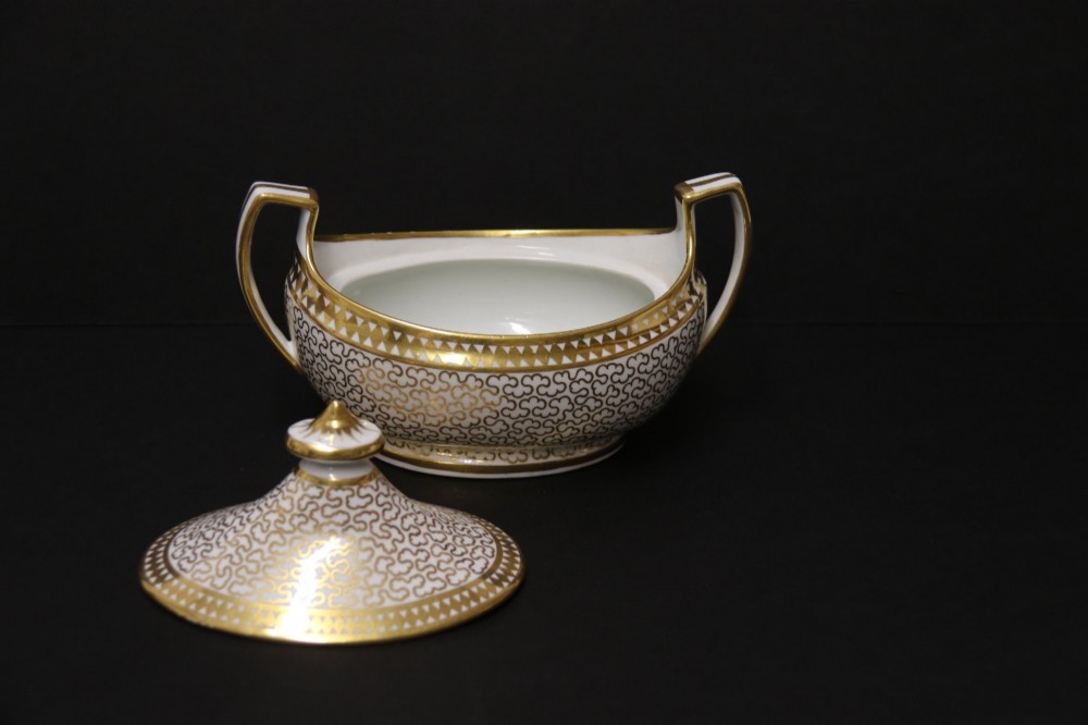 An antique white porcelain covered sucrier decorated with intricate gold scroll patterning, geometric trim, twin loop handles, and a pointed finial lid. View VI