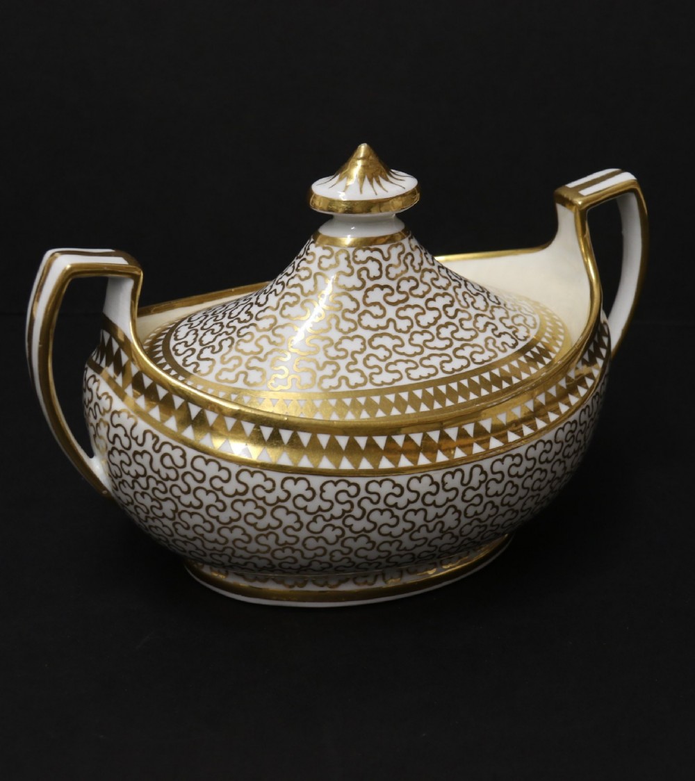 An antique white porcelain covered sucrier decorated with intricate gold scroll patterning, geometric trim, twin loop handles, and a pointed finial lid. View XIII