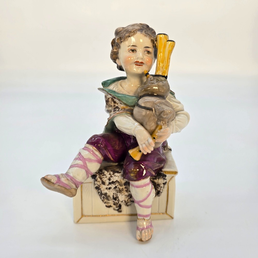 A 19th-century polychrome porcelain figurine depicting a seated boy playing bagpipes resting on a rectangular trunk with gilded trim. View II
