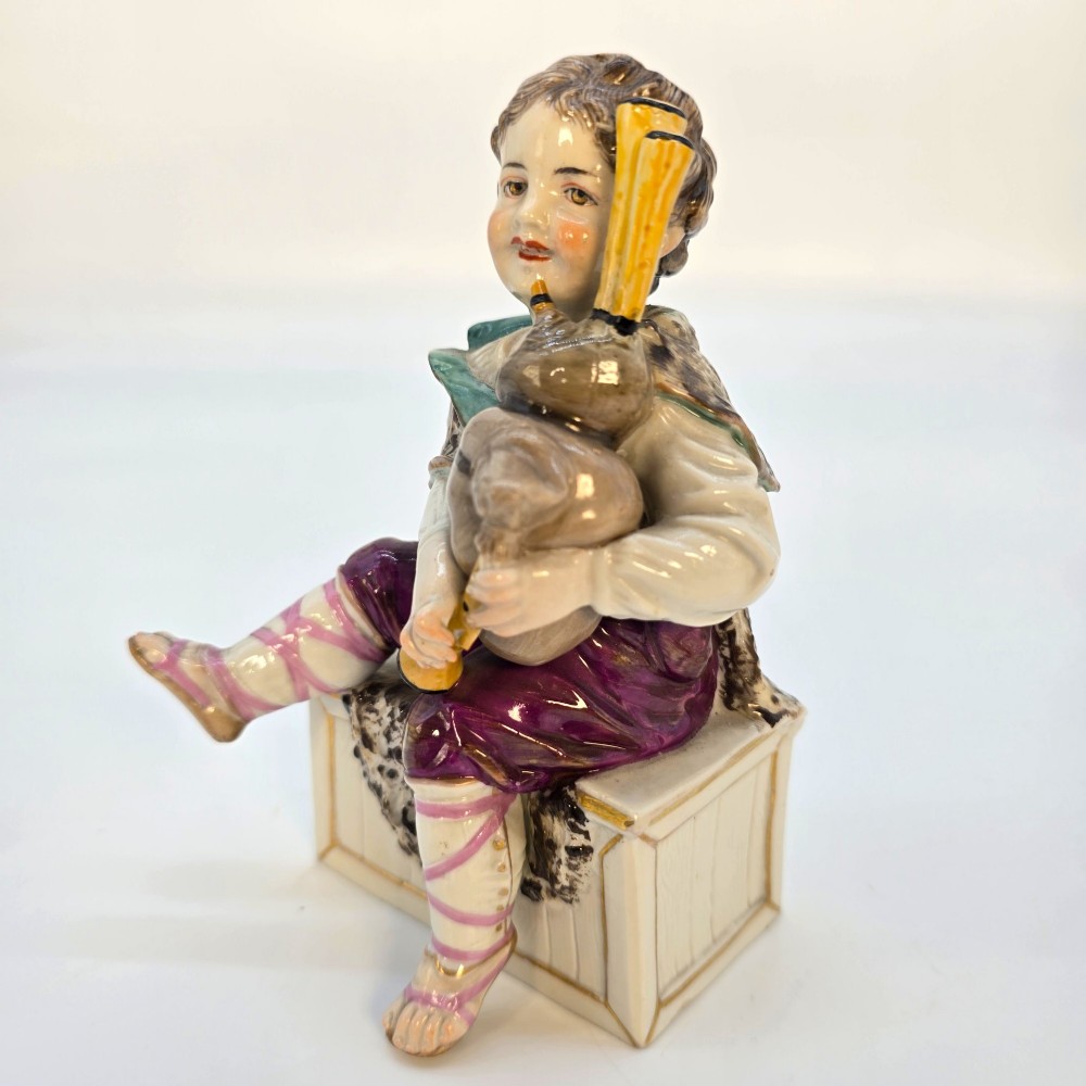 A 19th-century polychrome porcelain figurine depicting a seated boy playing bagpipes resting on a rectangular trunk with gilded trim. View III