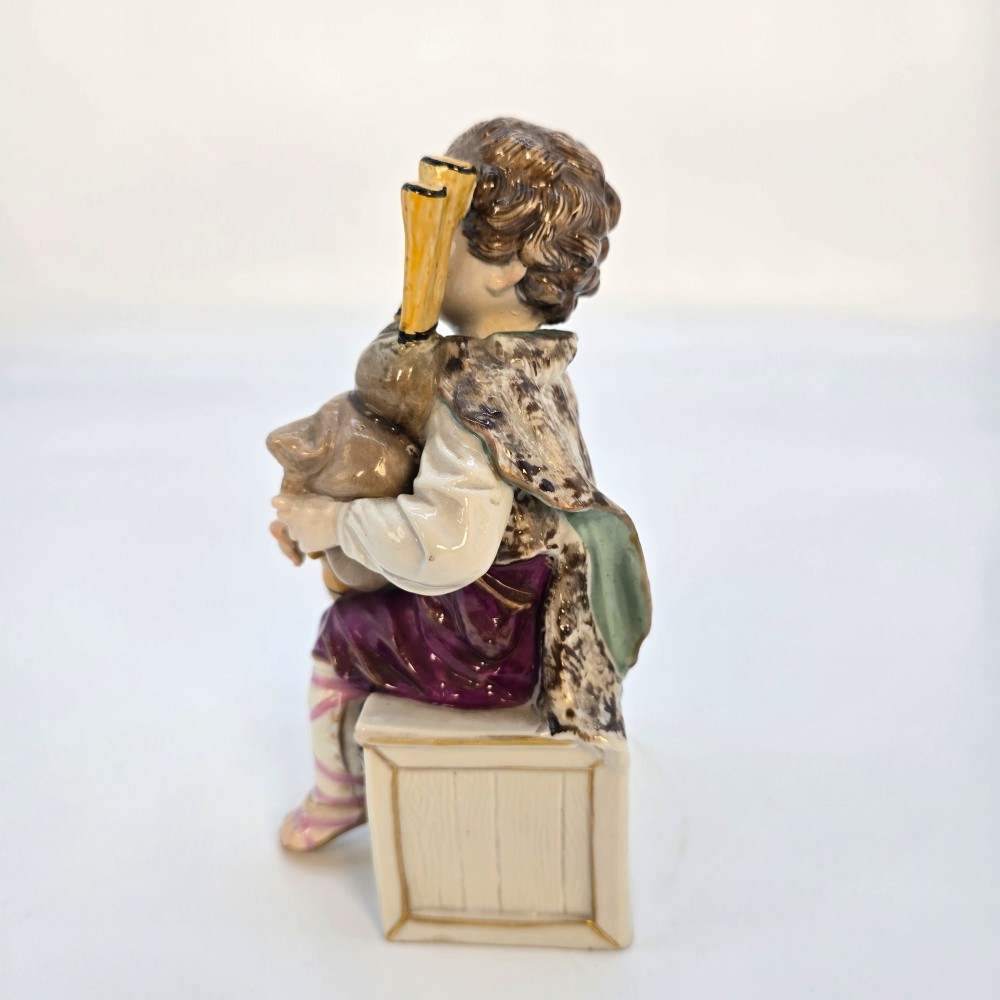 A 19th-century polychrome porcelain figurine depicting a seated boy playing bagpipes resting on a rectangular trunk with gilded trim. View IV