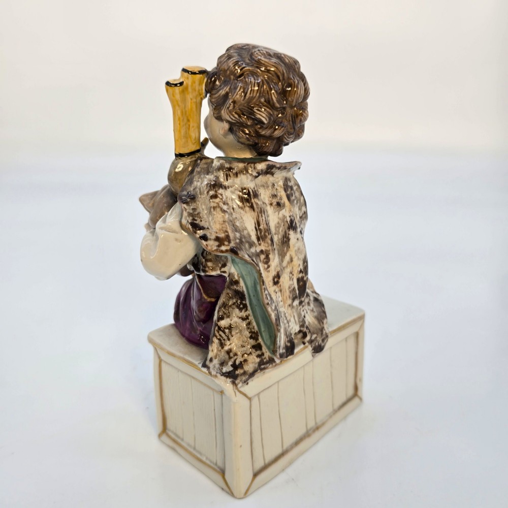 A 19th-century polychrome porcelain figurine depicting a seated boy playing bagpipes resting on a rectangular trunk with gilded trim. View V