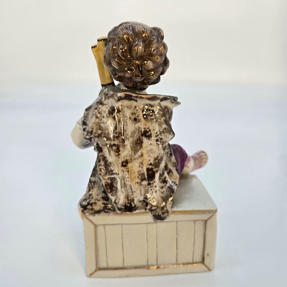 A 19th-century polychrome porcelain figurine depicting a seated boy playing bagpipes resting on a rectangular trunk with gilded trim. View VI