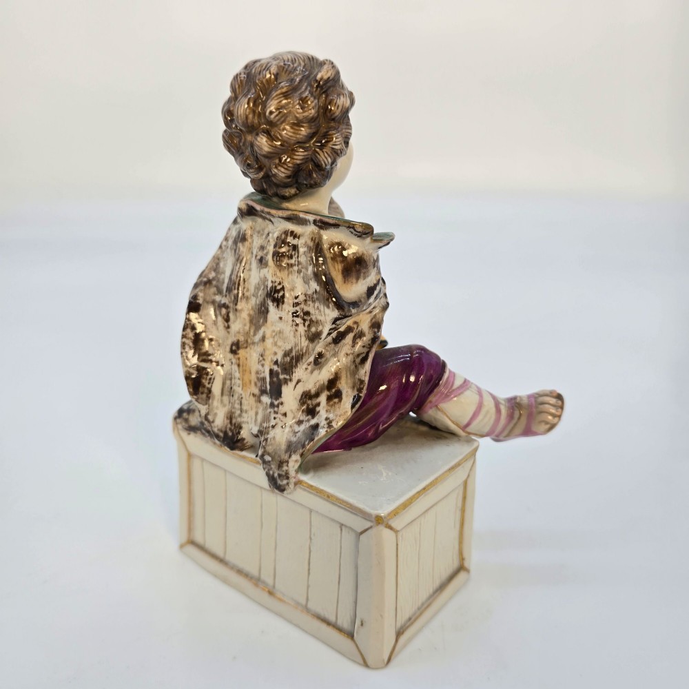 A 19th-century polychrome porcelain figurine depicting a seated boy playing bagpipes resting on a rectangular trunk with gilded trim. View VII