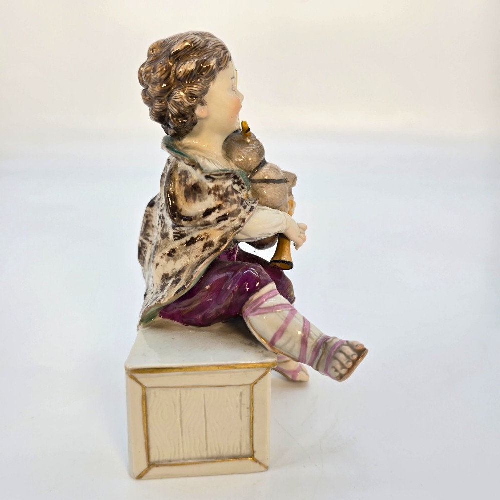 A 19th-century polychrome porcelain figurine depicting a seated boy playing bagpipes resting on a rectangular trunk with gilded trim. View VIII