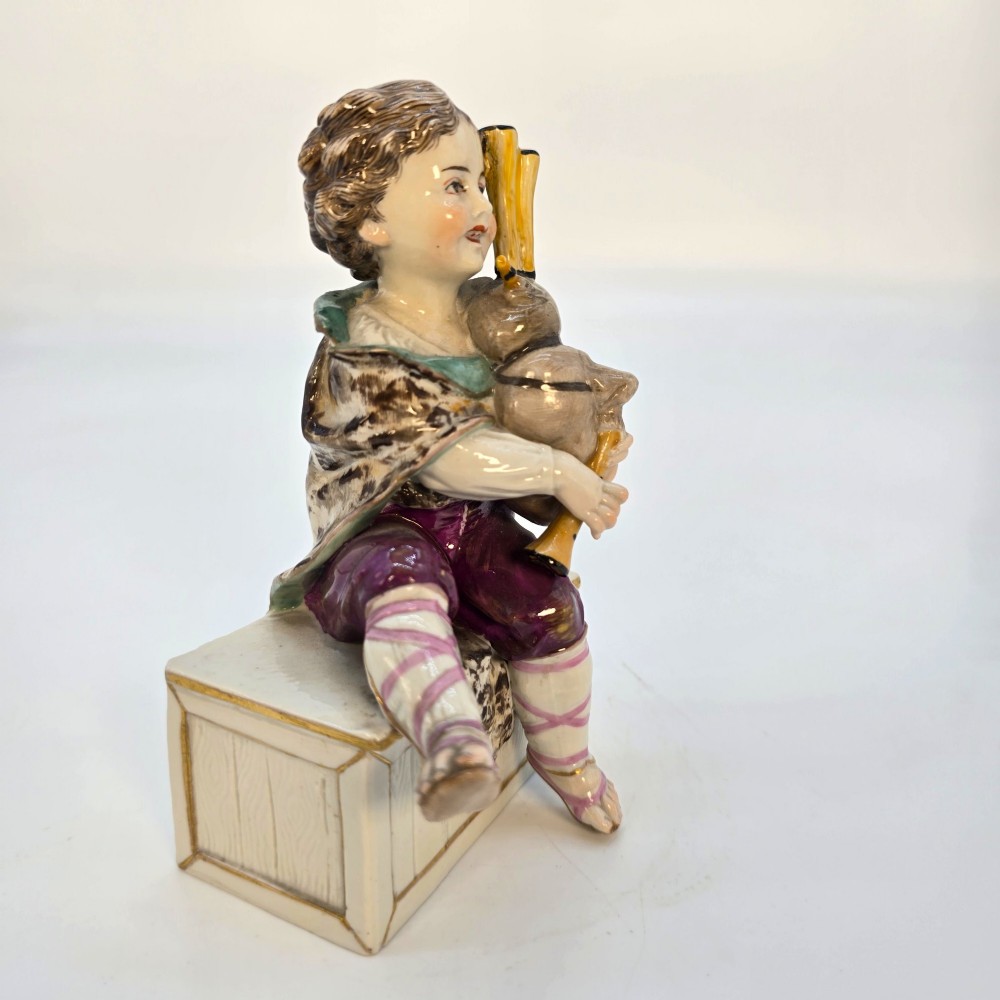 A 19th-century polychrome porcelain figurine depicting a seated boy playing bagpipes resting on a rectangular trunk with gilded trim. View IX