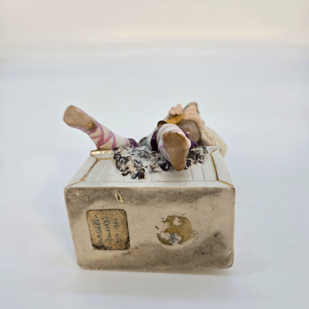 A 19th-century polychrome porcelain figurine depicting a seated boy playing bagpipes resting on a rectangular trunk with gilded trim. View X