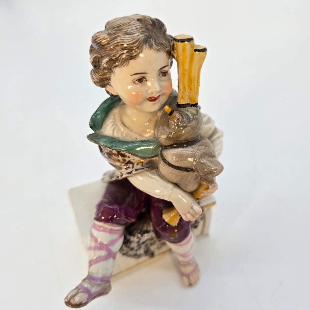 A 19th-century polychrome porcelain figurine depicting a seated boy playing bagpipes resting on a rectangular trunk with gilded trim. View XI