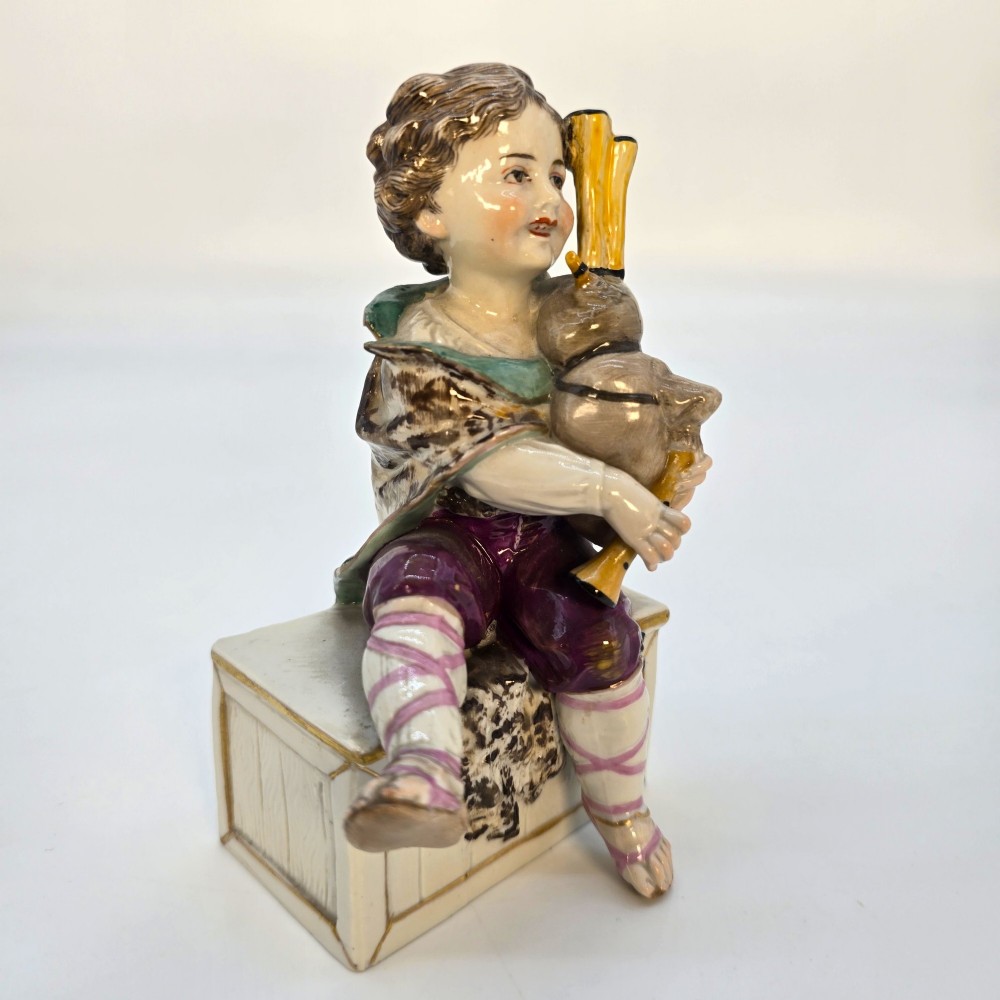 A 19th-century polychrome porcelain figurine depicting a seated boy playing bagpipes resting on a rectangular trunk with gilded trim. View XII