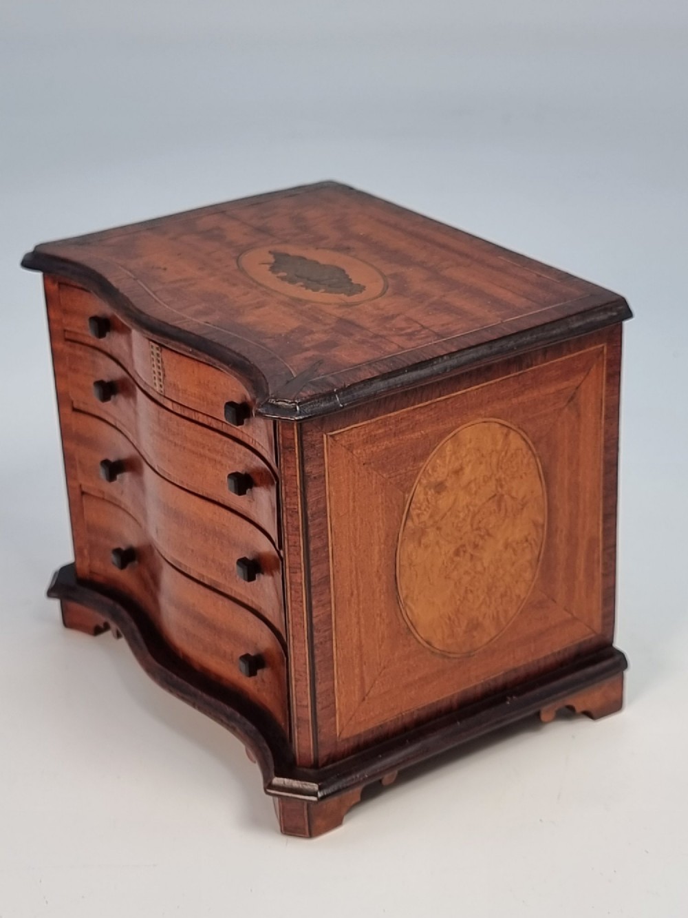 An Edwardian miniature satinwood chest of drawers featuring a serpentine front, inlaid shell medallion on the top, and four small drawers with wooden knobs. View VI
