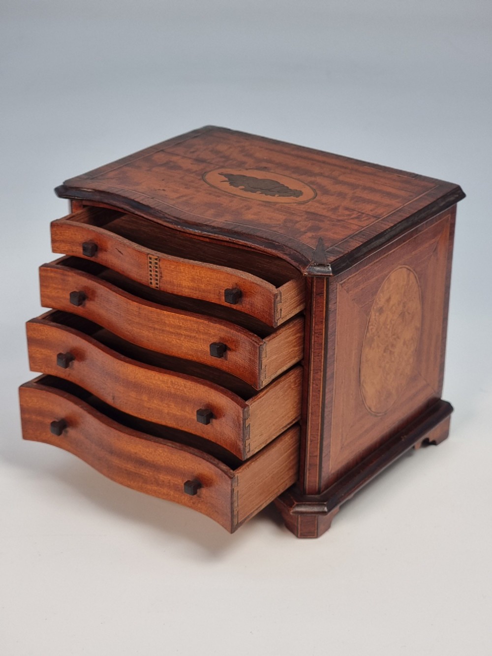 An Edwardian miniature satinwood chest of drawers featuring a serpentine front, inlaid shell medallion on the top, and four small drawers with wooden knobs. View VII
