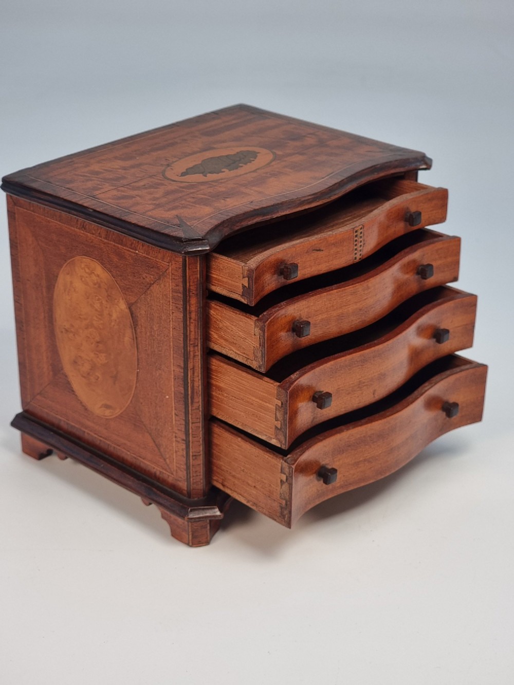 An Edwardian miniature satinwood chest of drawers featuring a serpentine front, inlaid shell medallion on the top, and four small drawers with wooden knobs. View VIII
