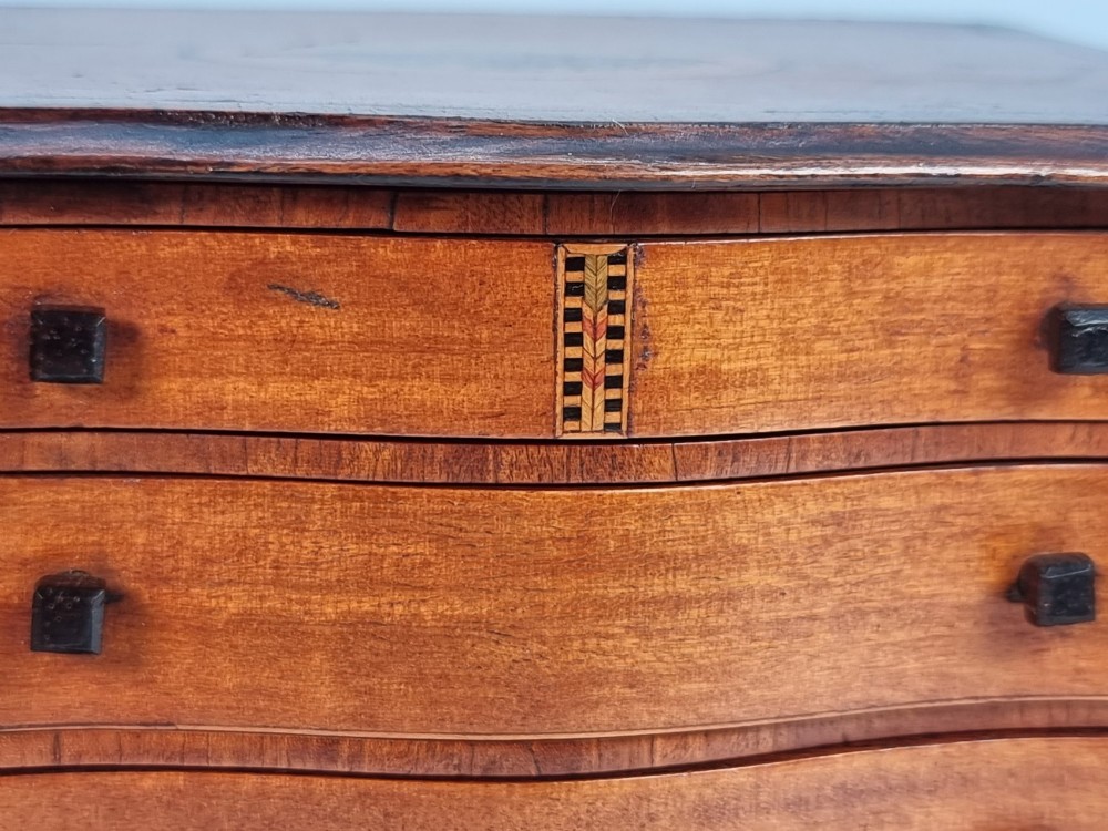 An Edwardian miniature satinwood chest of drawers featuring a serpentine front, inlaid shell medallion on the top, and four small drawers with wooden knobs. View X