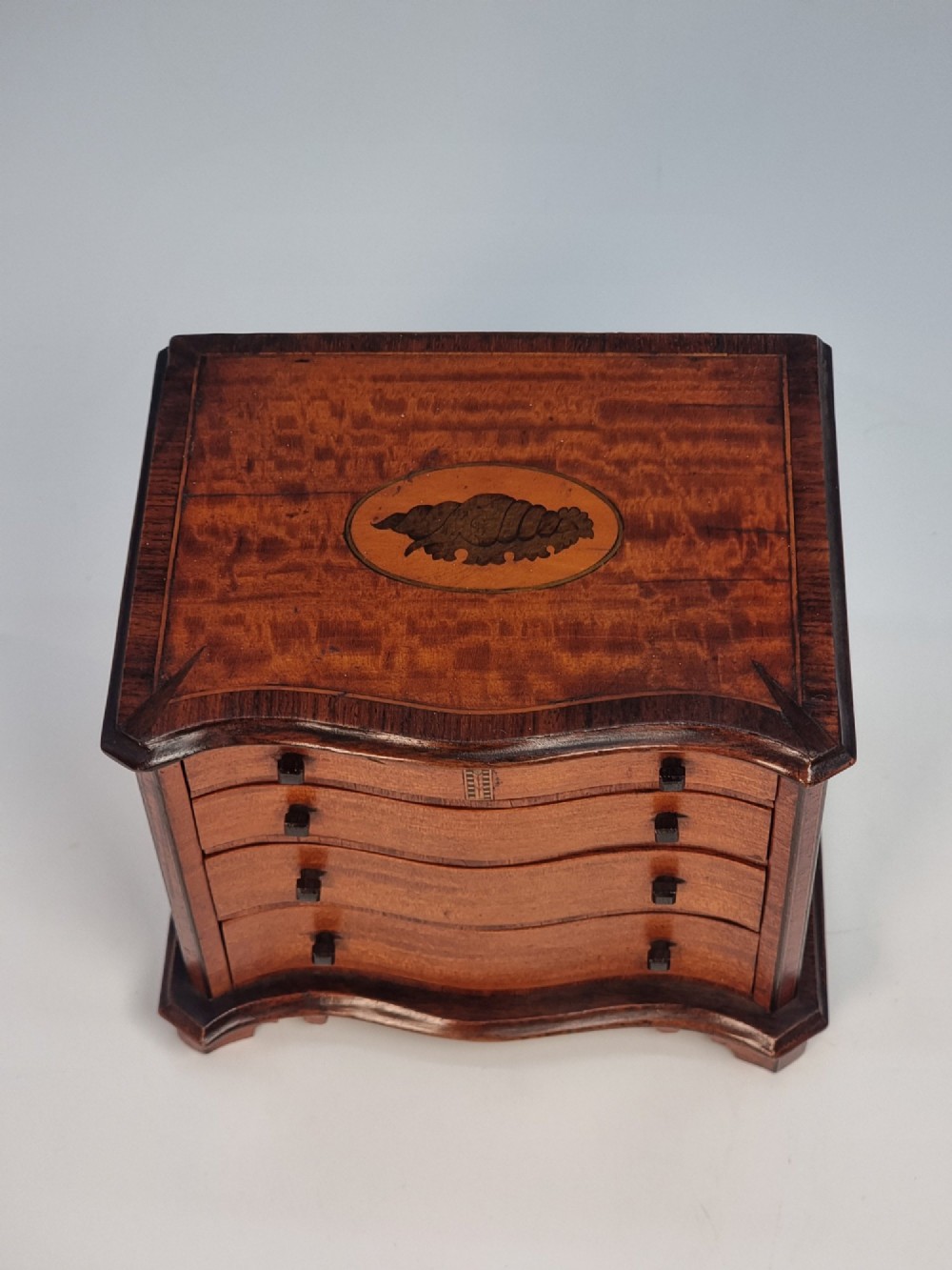 An Edwardian miniature satinwood chest of drawers featuring a serpentine front, inlaid shell medallion on the top, and four small drawers with wooden knobs. View XII