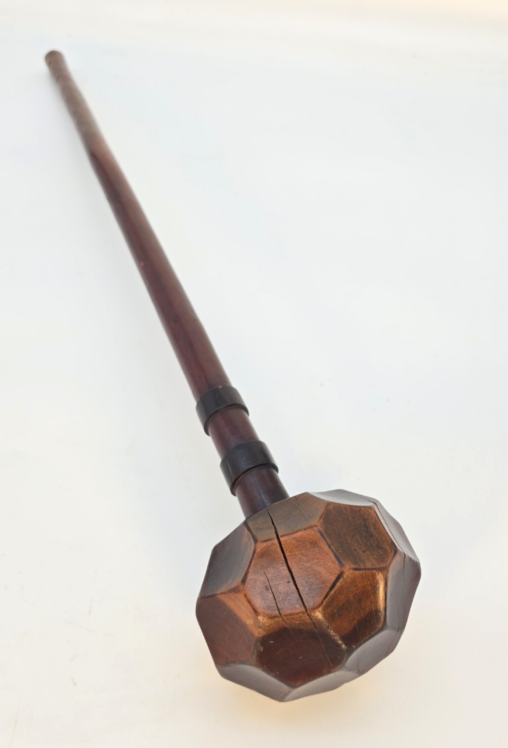 An antique dark hardwood Zulu knobkerrie club featuring a faceted spherical head and a slender shaft with raised carved rings near the top. View II