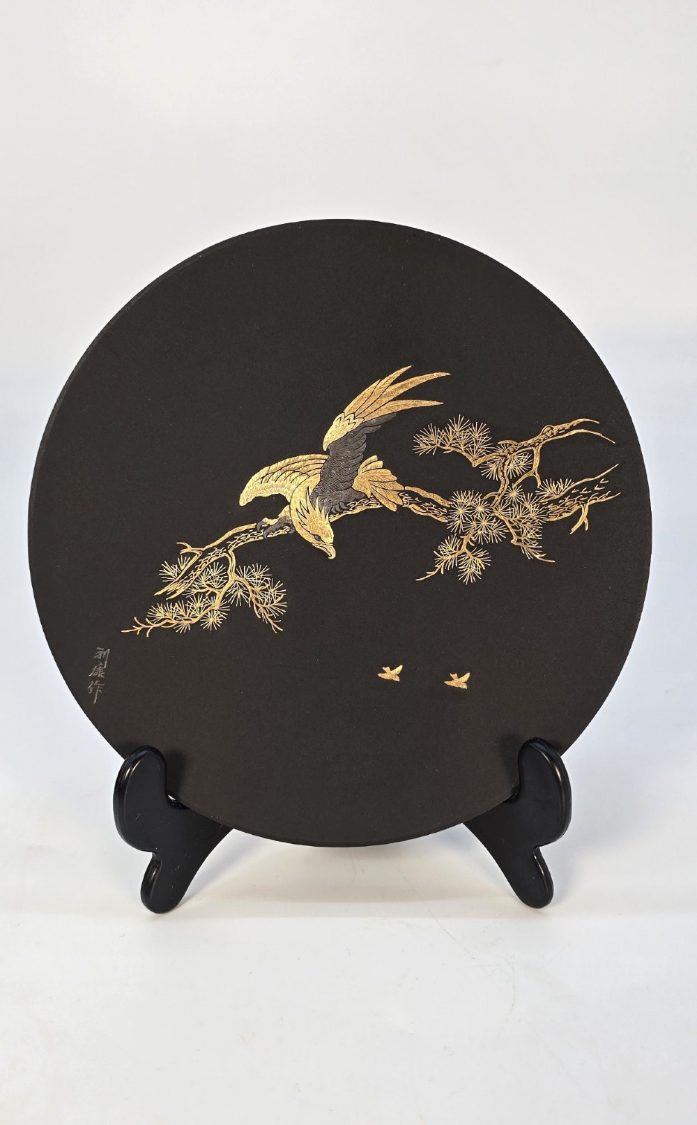 A circular black patinated iron plaque featuring an inlaid gold and silver depiction of an eagle perched on a pine branch.