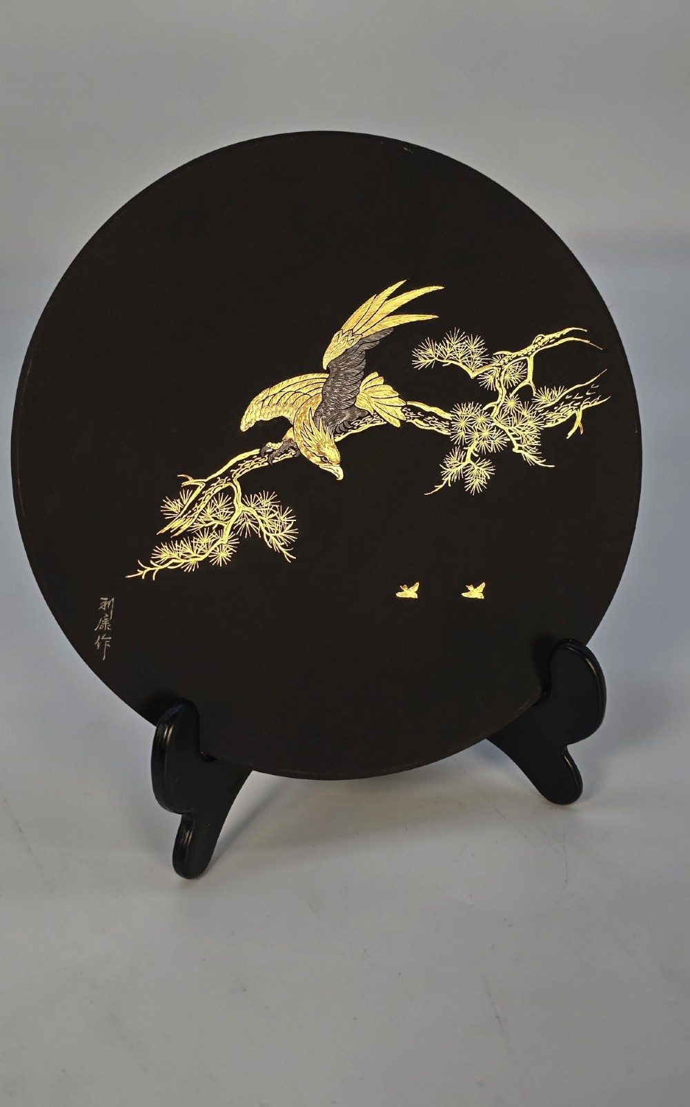 A circular black patinated iron plaque featuring an inlaid gold and silver depiction of an eagle perched on a pine branch. View II