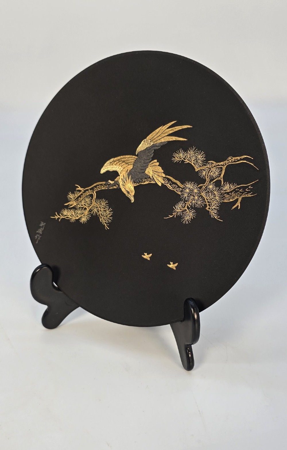 A circular black patinated iron plaque featuring an inlaid gold and silver depiction of an eagle perched on a pine branch. View III