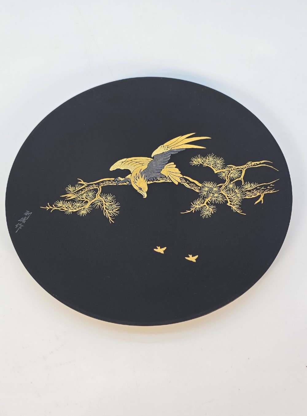 A circular black patinated iron plaque featuring an inlaid gold and silver depiction of an eagle perched on a pine branch. View IX