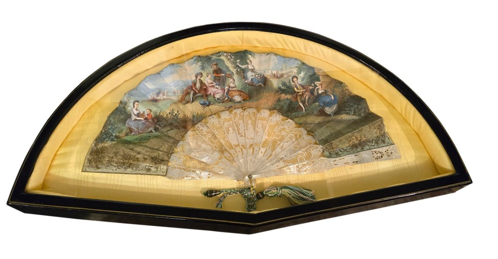 A late 18th-century Italian folding fan featuring a hand-painted pastoral scene mounted on carved mother-of-pearl sticks, presented inside a fan-shaped black frame with a cream silk lining.