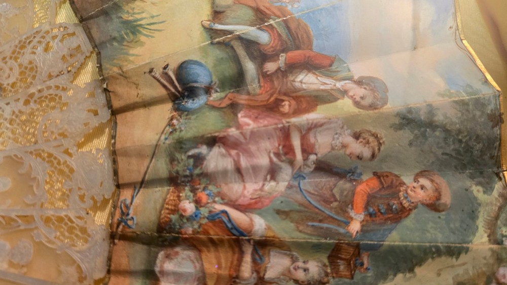 A late 18th-century Italian folding fan featuring a hand-painted pastoral scene mounted on carved mother-of-pearl sticks, presented inside a fan-shaped black frame with a cream silk lining. View II