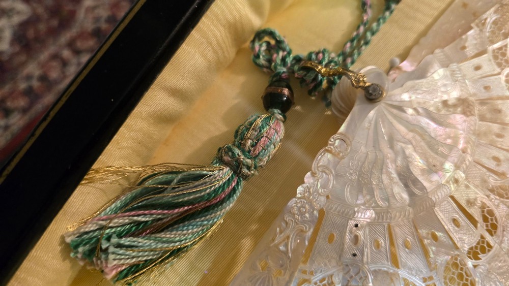 A late 18th-century Italian folding fan featuring a hand-painted pastoral scene mounted on carved mother-of-pearl sticks, presented inside a fan-shaped black frame with a cream silk lining. View III