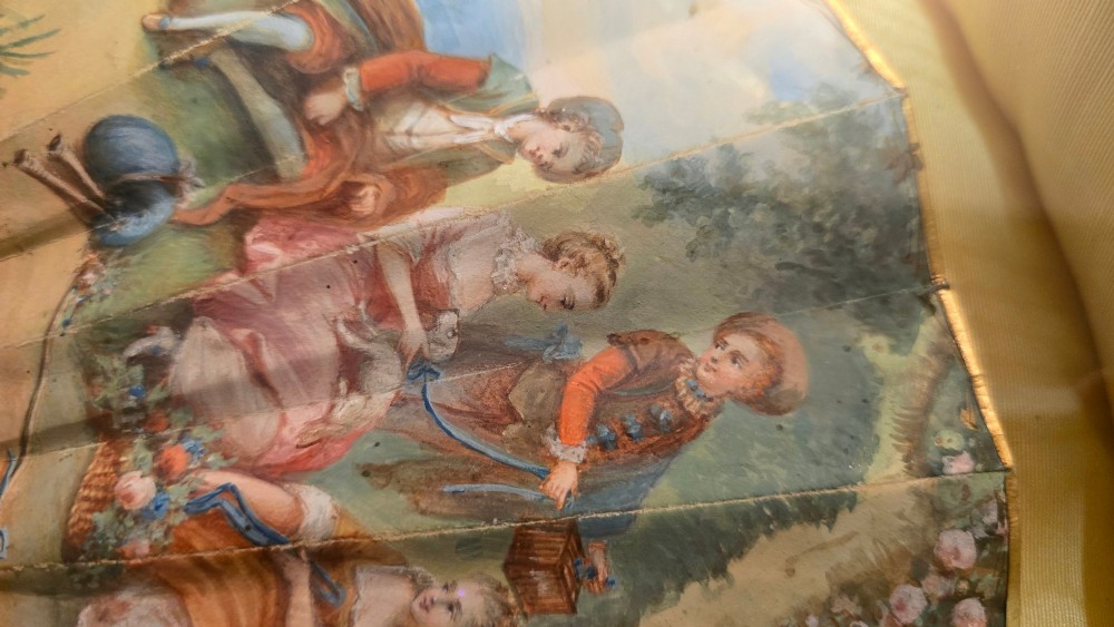 A late 18th-century Italian folding fan featuring a hand-painted pastoral scene mounted on carved mother-of-pearl sticks, presented inside a fan-shaped black frame with a cream silk lining. View IV