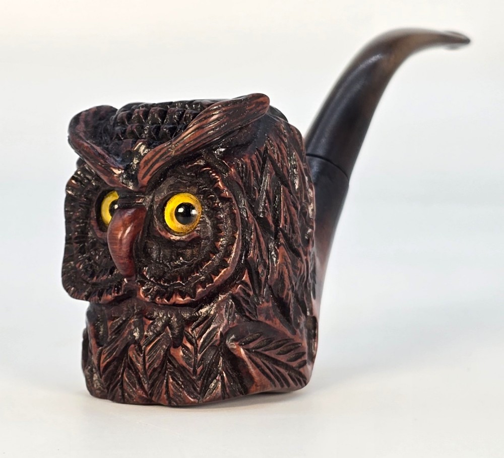 A detailed hand-carved briar wood tobacco pipe crafted in the shape of an owls head featuring yellow glass eyes and a curved black stem.