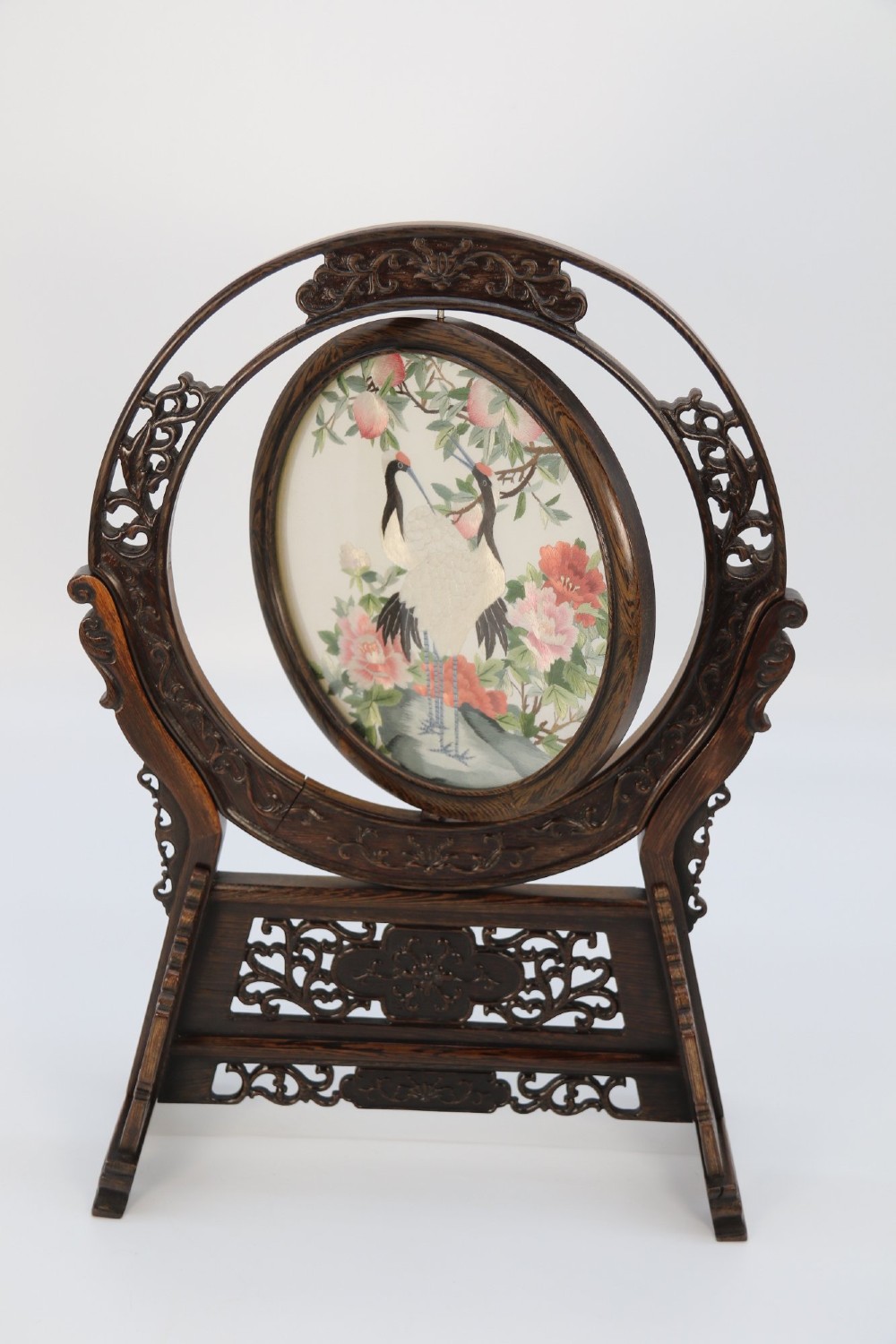 A circular double-sided silk embroidery depicting cranes among pink flowers, set within a rotating carved dark hardwood frame and stand. View V