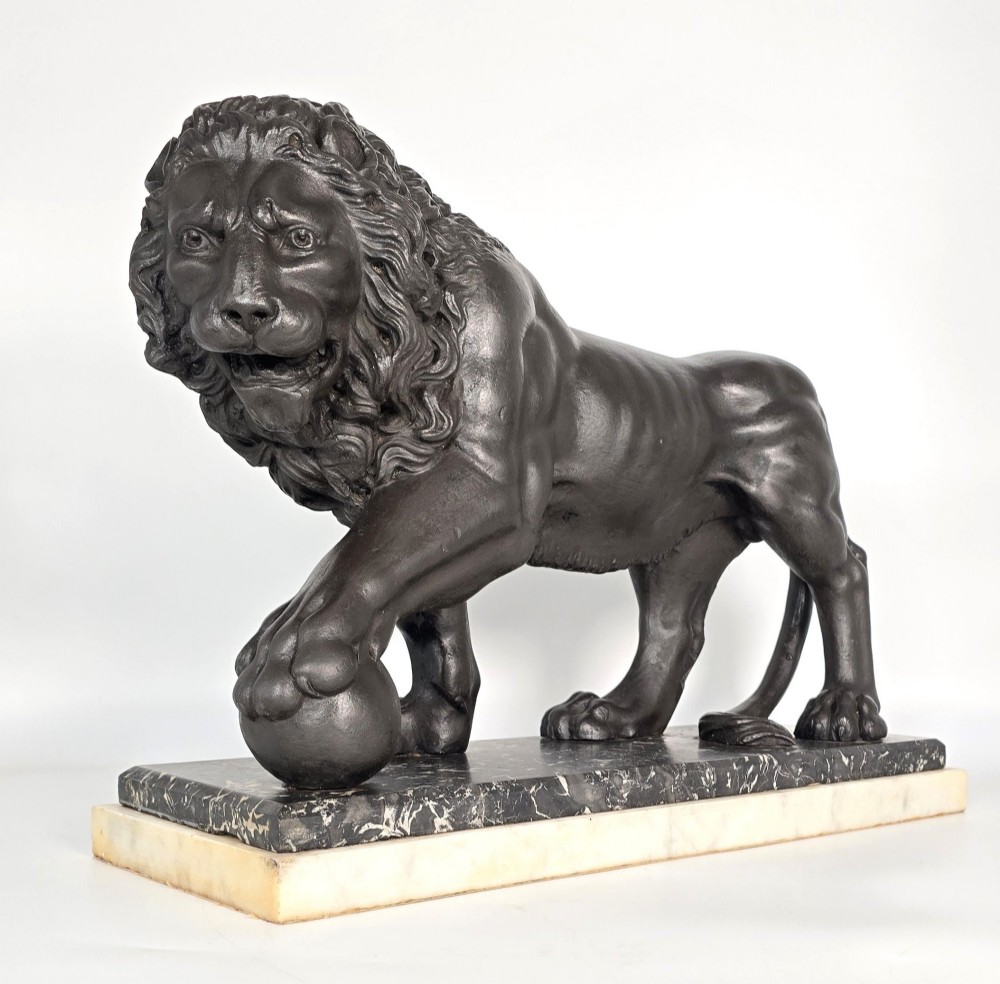 A dark patinated bronze sculpture of a lion resting its paw on a sphere, mounted on a two-tier marble plinth.