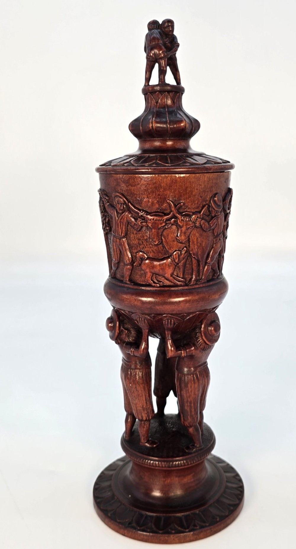 A tall hand-carved dark walnut lidded chalice featuring a stem sculpted as human figures, a relief carving of cattle around the vessel, and a finial with carved figures. View V