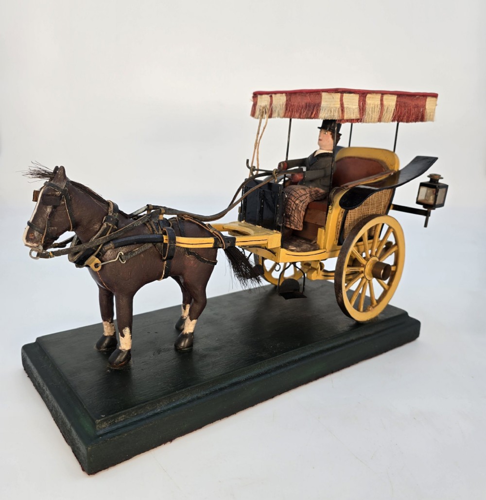 A hand-crafted 1928 wooden folk art model featuring a brown horse attached to a yellow two-wheeled chaise carriage with a driver figure under a striped canopy, mounted on a dark wooden base.