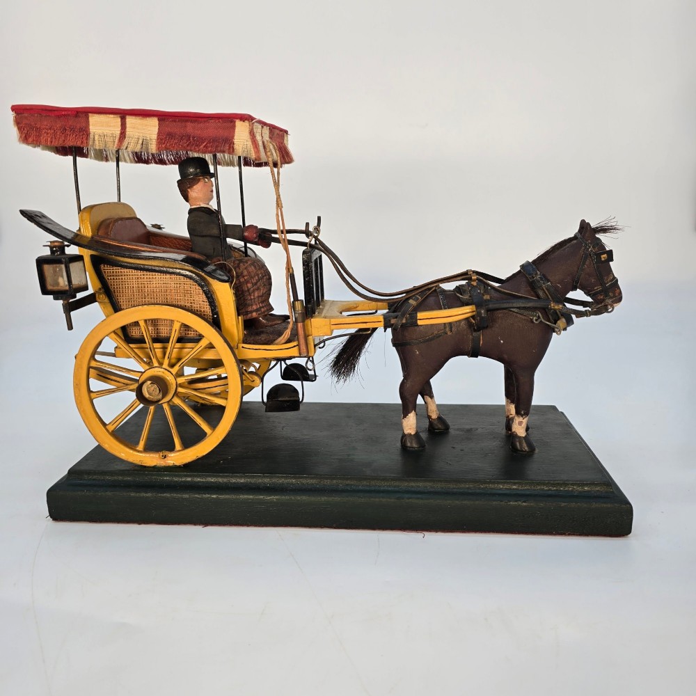 A hand-crafted 1928 wooden folk art model featuring a brown horse attached to a yellow two-wheeled chaise carriage with a driver figure under a striped canopy, mounted on a dark wooden base. View II