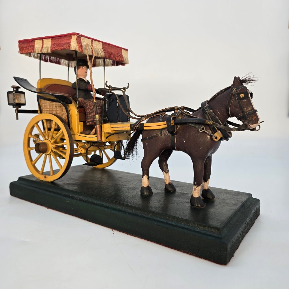 A hand-crafted 1928 wooden folk art model featuring a brown horse attached to a yellow two-wheeled chaise carriage with a driver figure under a striped canopy, mounted on a dark wooden base. View III